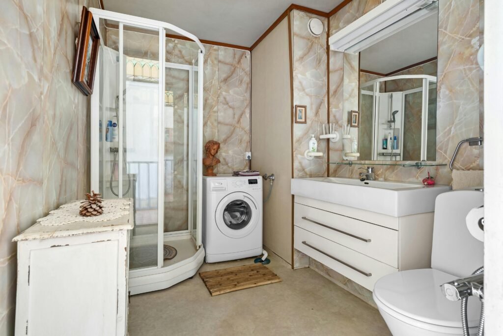 A bright, minimalist bathroom featuring a shower, sink, and washing machine setup.